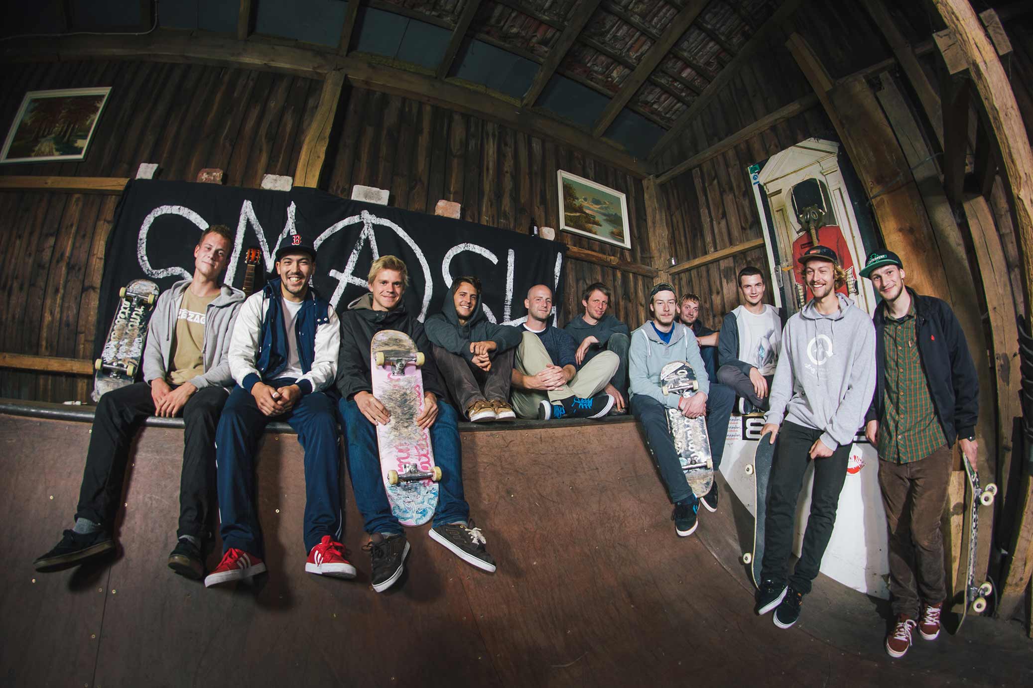 A group of 11 smiling young men are sitting or standing around a skateboard ramp in a barn-like building. Some of the men are holding skateboards.